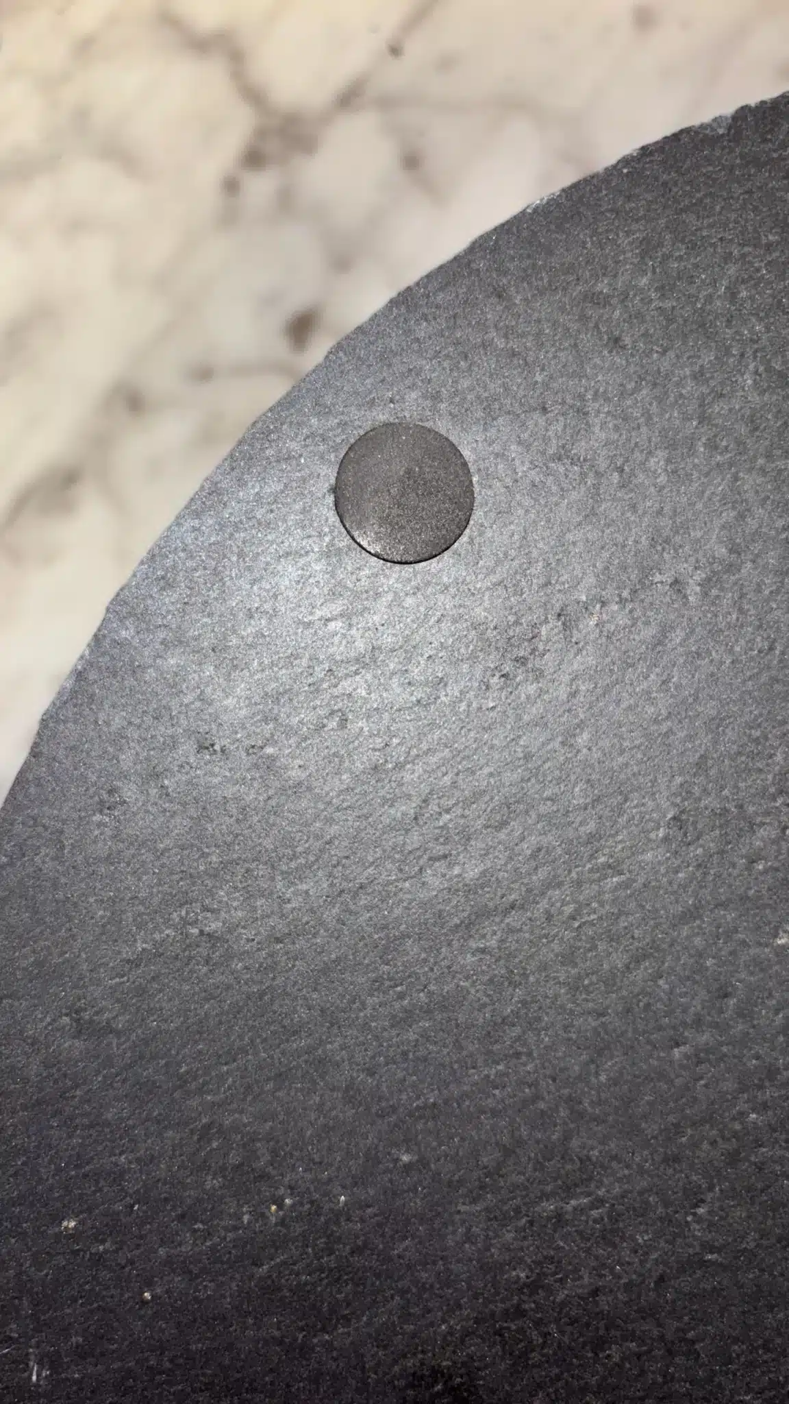 Close-up of a small round magnet attached to the edge of a dark, textured circular surface, possibly a slate board, with a blurred white and gray background.