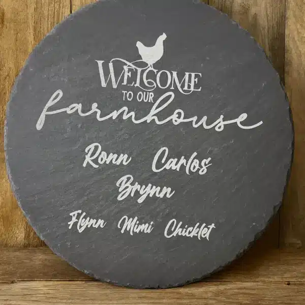 A round slate sign with white text reads “Welcome to our farmhouse” with a chicken graphic above. Below are the names: Ronn, Carlos, Brynn, Flynn, Mimi, and Chicklet. The sign is displayed against a wooden background.