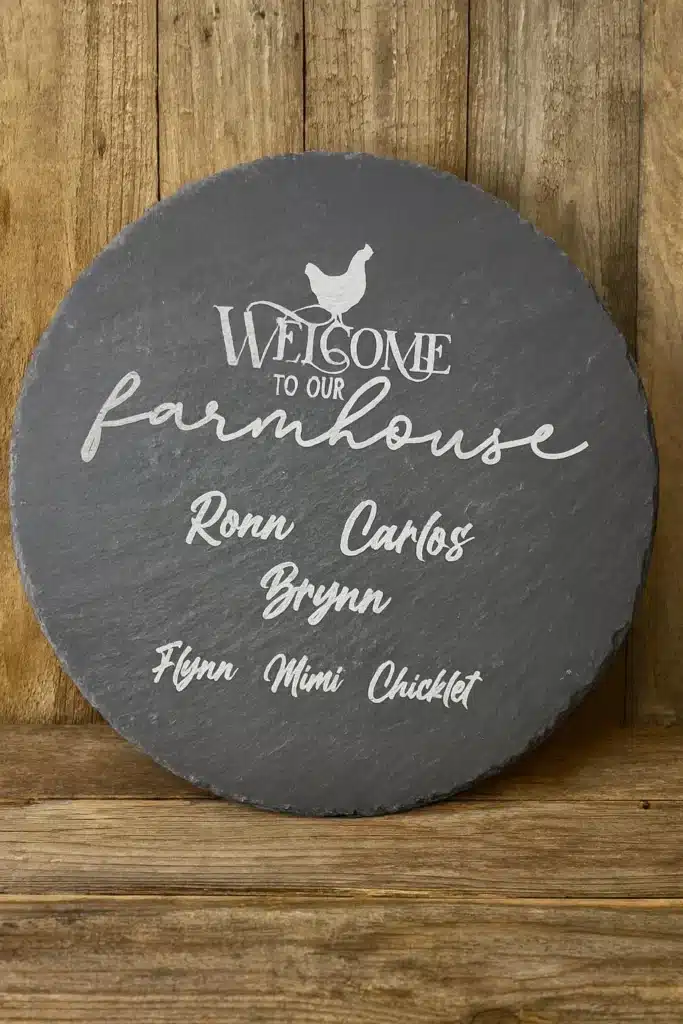 A round slate sign with white text reads “Welcome to our farmhouse” with a chicken graphic above. Below are the names: Ronn, Carlos, Brynn, Flynn, Mimi, and Chicklet. The sign is displayed against a wooden background.