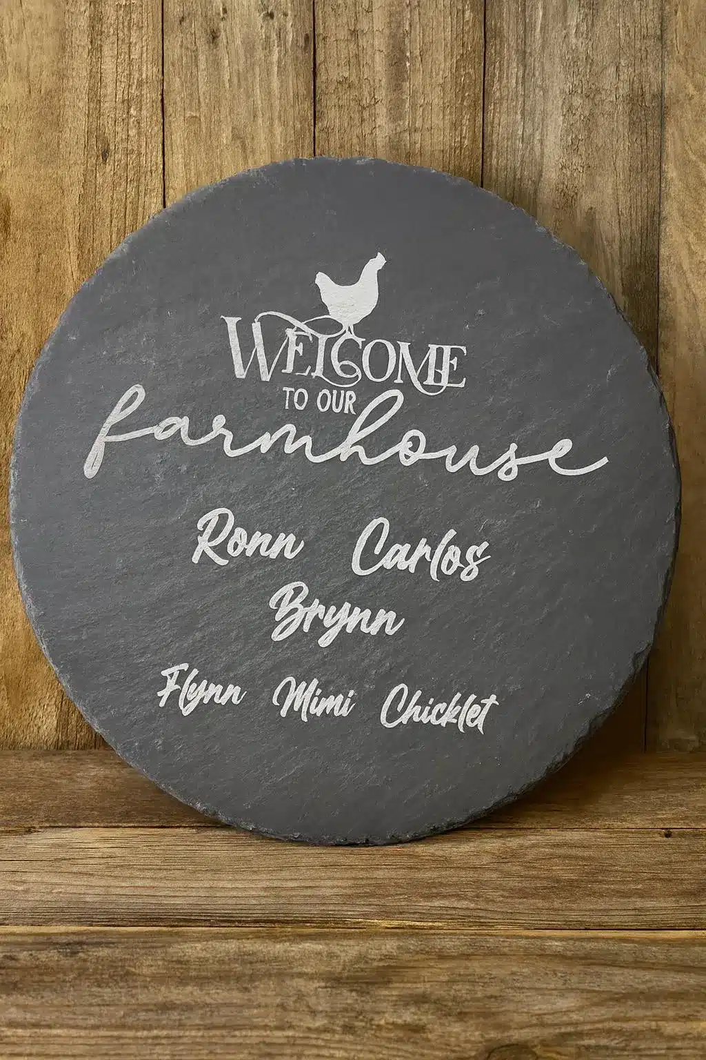 A round slate sign with white text reads “Welcome to our farmhouse” with a chicken graphic above. Below are the names: Ronn, Carlos, Brynn, Flynn, Mimi, and Chicklet. The sign is displayed against a wooden background.