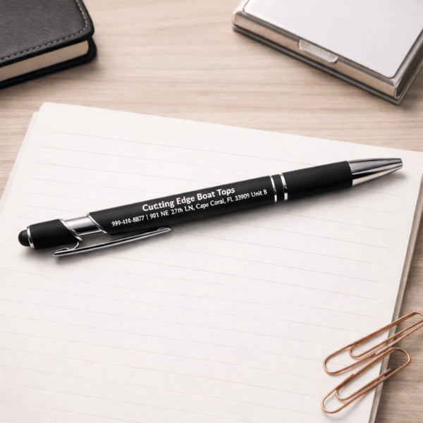 A Rubberized Metal Ballpoint Pen with Stylus Tip featuring white text rests on a blank, lined notebook page beside paperclips, a closed notebook, and a silver cardholder atop a wooden desk.
