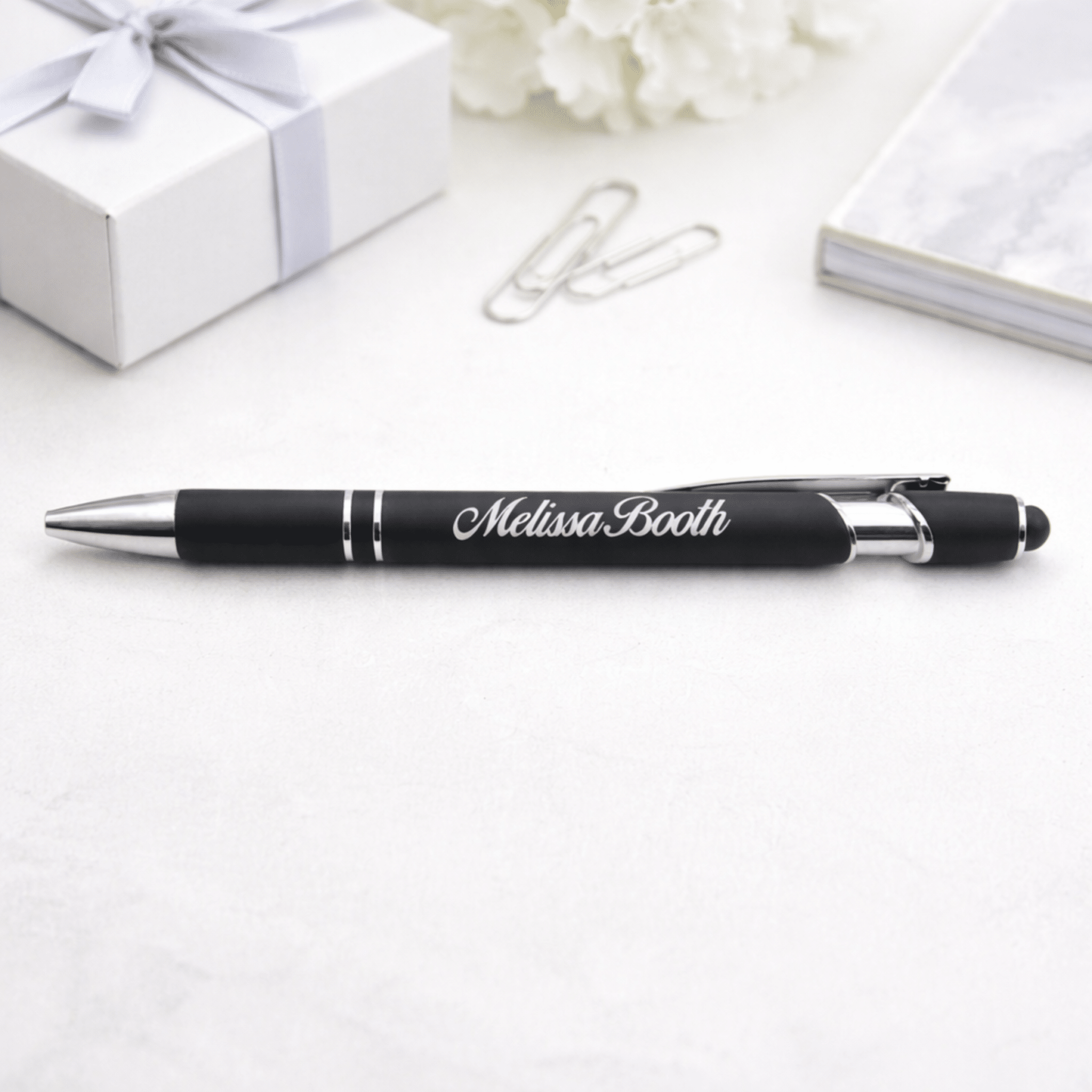 A black and silver Rubberized Metal Ballpoint Pen with Stylus Tip, engraved with Melissa Booth, rests on a white surface near a silver gift box, two paper clips, and a marble-patterned notebook.