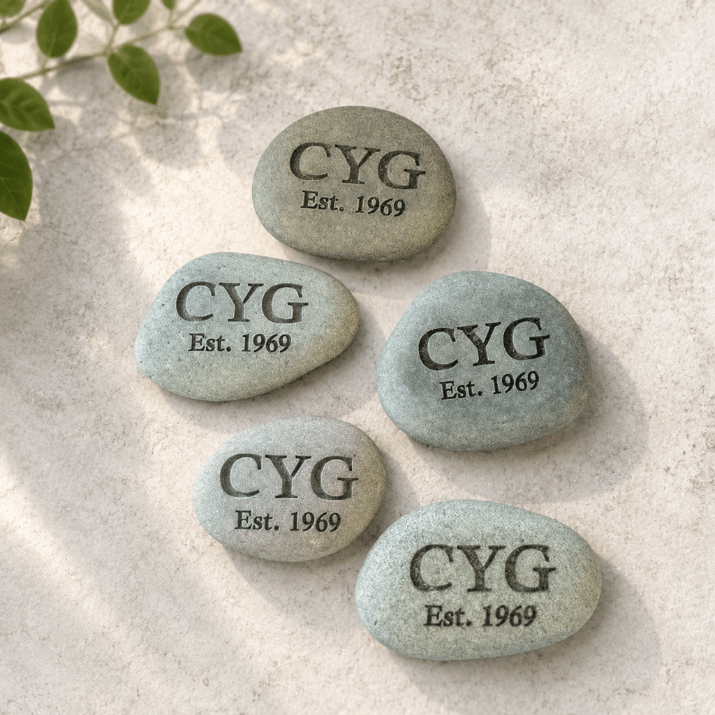 Five smooth stones on a light surface, each engraved with CYG Est. 1969. Sunlight and shadows fall across the scene, with green leaves in the upper left corner.