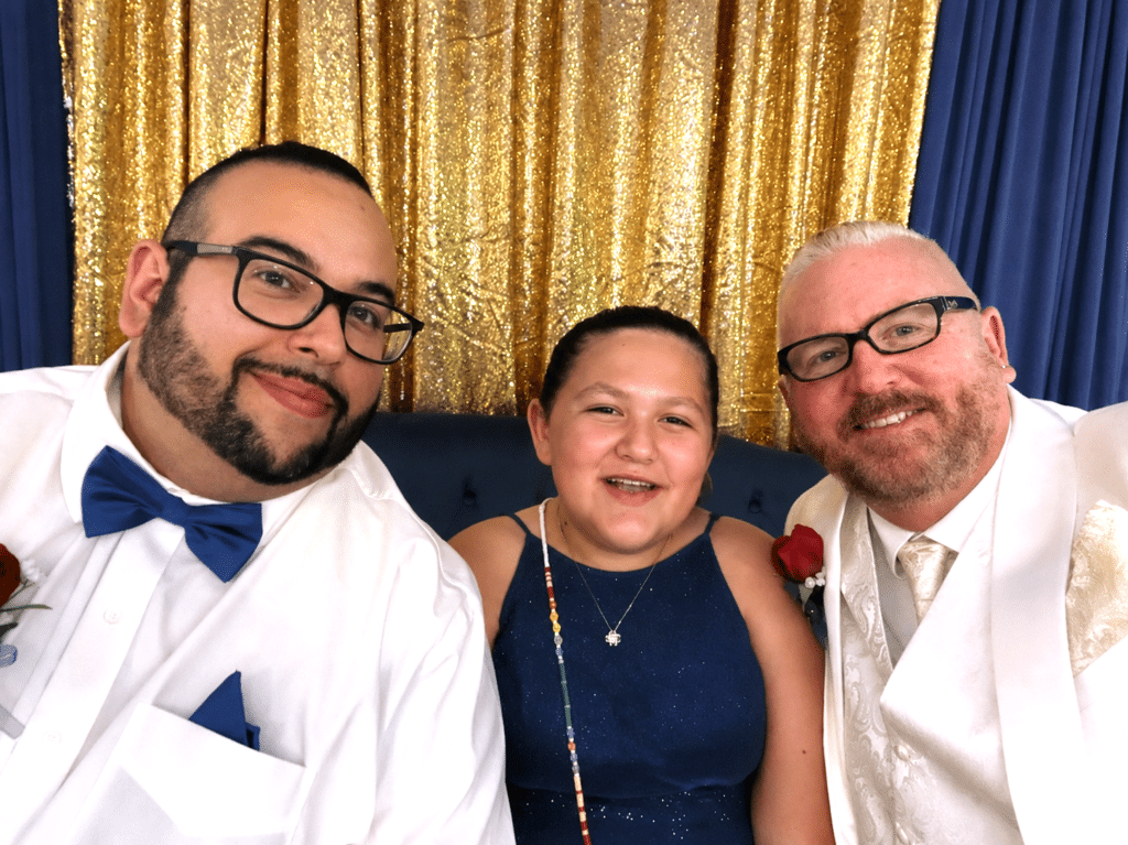 72b0d1e6-e452-4c3b-a6ab-f28dae53ba99 Three people dressed formally, smiling and sitting together in front of a gold sequin curtain. The two men wear glasses and suits; the girl in the center wears a navy blue dress with a jeweled necklace.