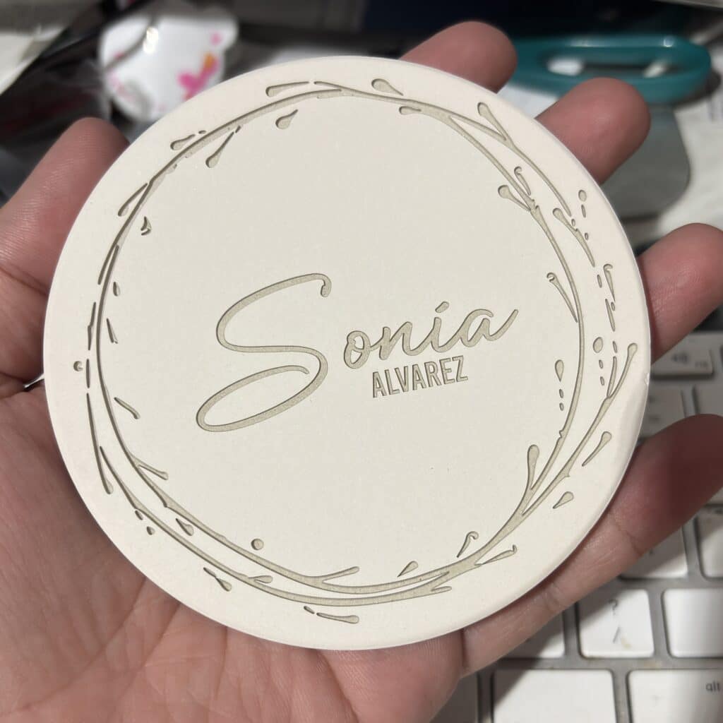 A hand holds a round, off-white card with Sonia Alvarez embossed in elegant script, surrounded by a decorative leafy border. The background includes a keyboard and office supplies.