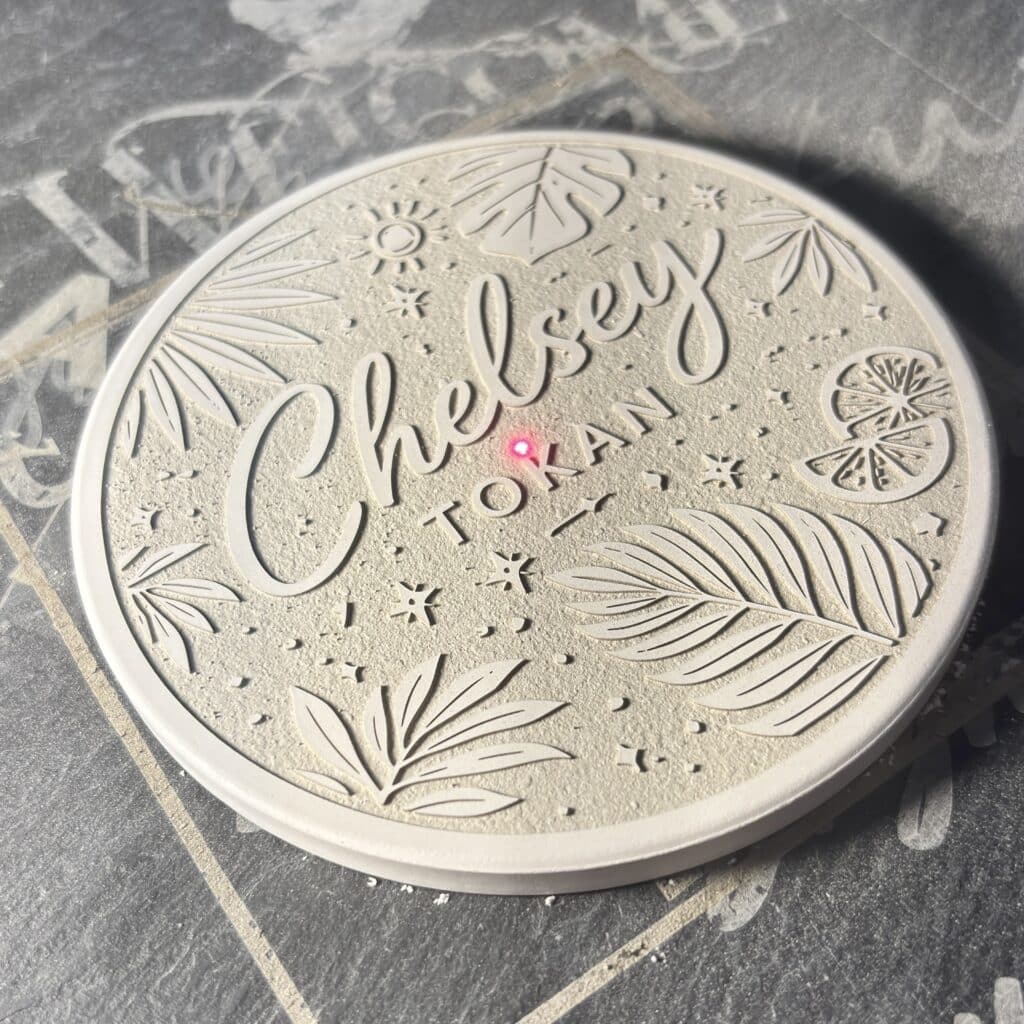 A round, textured coaster with tropical leaf and citrus slice designs, engraved with “Chelsey Jordan.” A small red laser dot is visible on the coaster’s surface. The background is a gray, chalky surface.