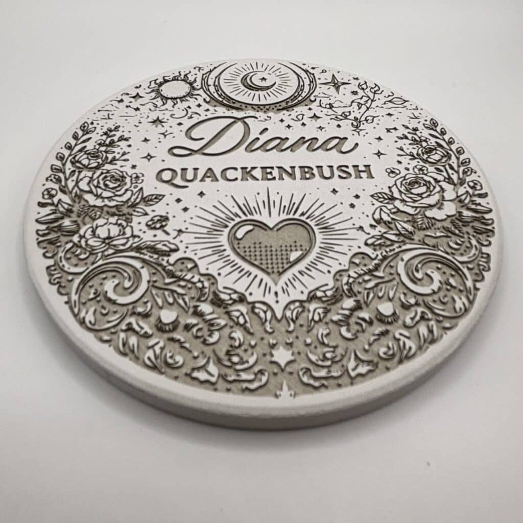 A round, engraved stone coaster with floral designs, a heart, stars, and the words Diana QUACKENBUSH in the center on a white background.