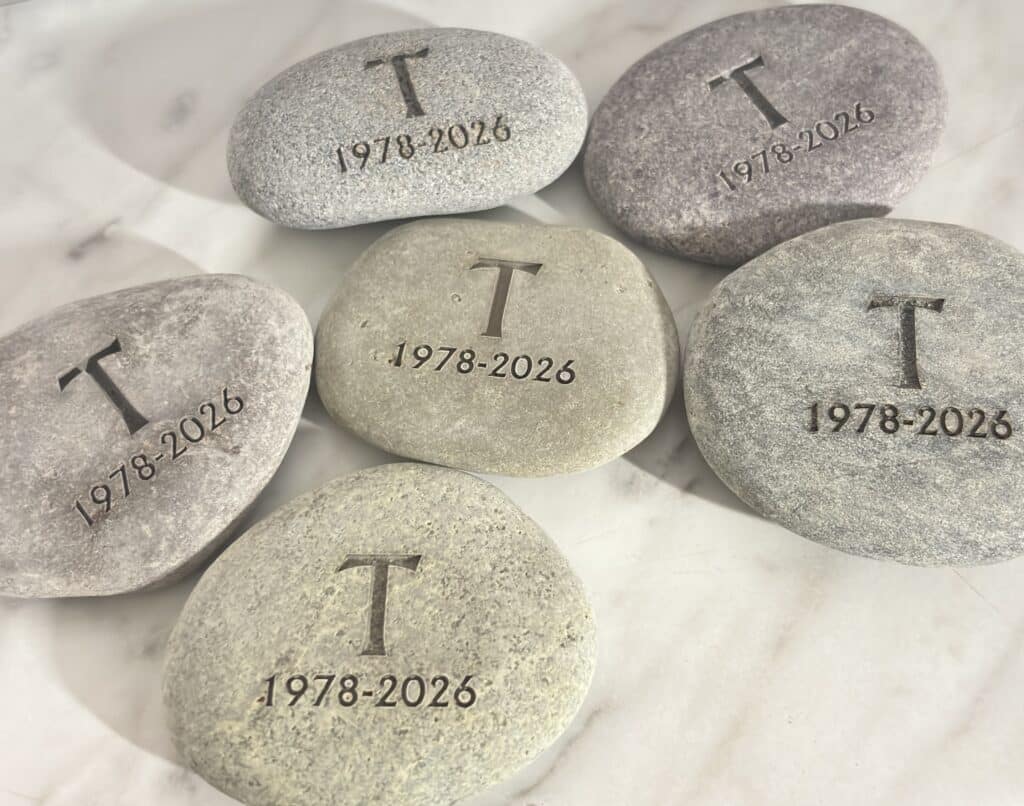 Six engraved stones on a light surface, each marked with the letter T and the years 1978-2026. The stones vary in color from gray to beige.