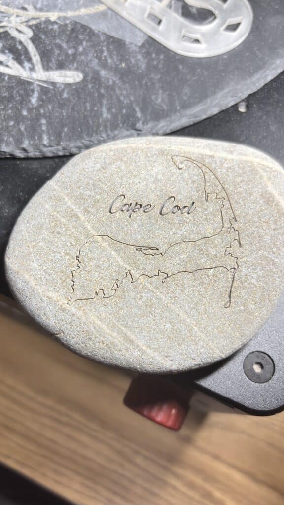 A smooth rock engraved with an outline map of Cape Cod and the words Cape Cod, placed on a black surface with part of another object visible in the background.