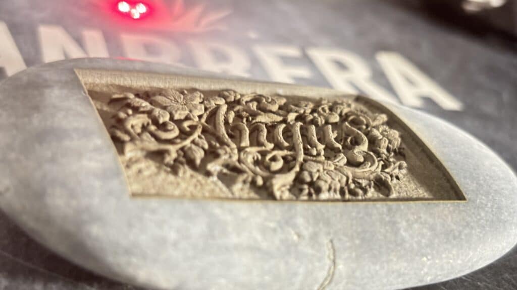 A close-up of a decorative metal plaque with ornate floral carvings and the word August engraved, resting on a smooth stone. Blurred white letters and a red light are visible in the background.