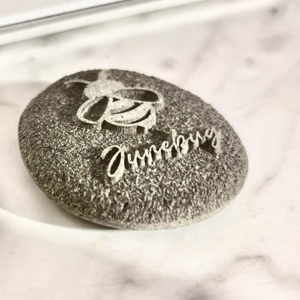 A textured stone features an engraved cartoon bee above the word Dumpling, both carved into its surface. The stone rests on a white marble background.