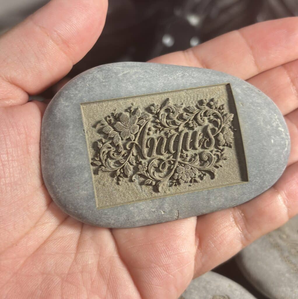 A hand holds a smooth stone with an ornate floral engraving and the word Angus in the center. Sunlight highlights the intricate details of the design.