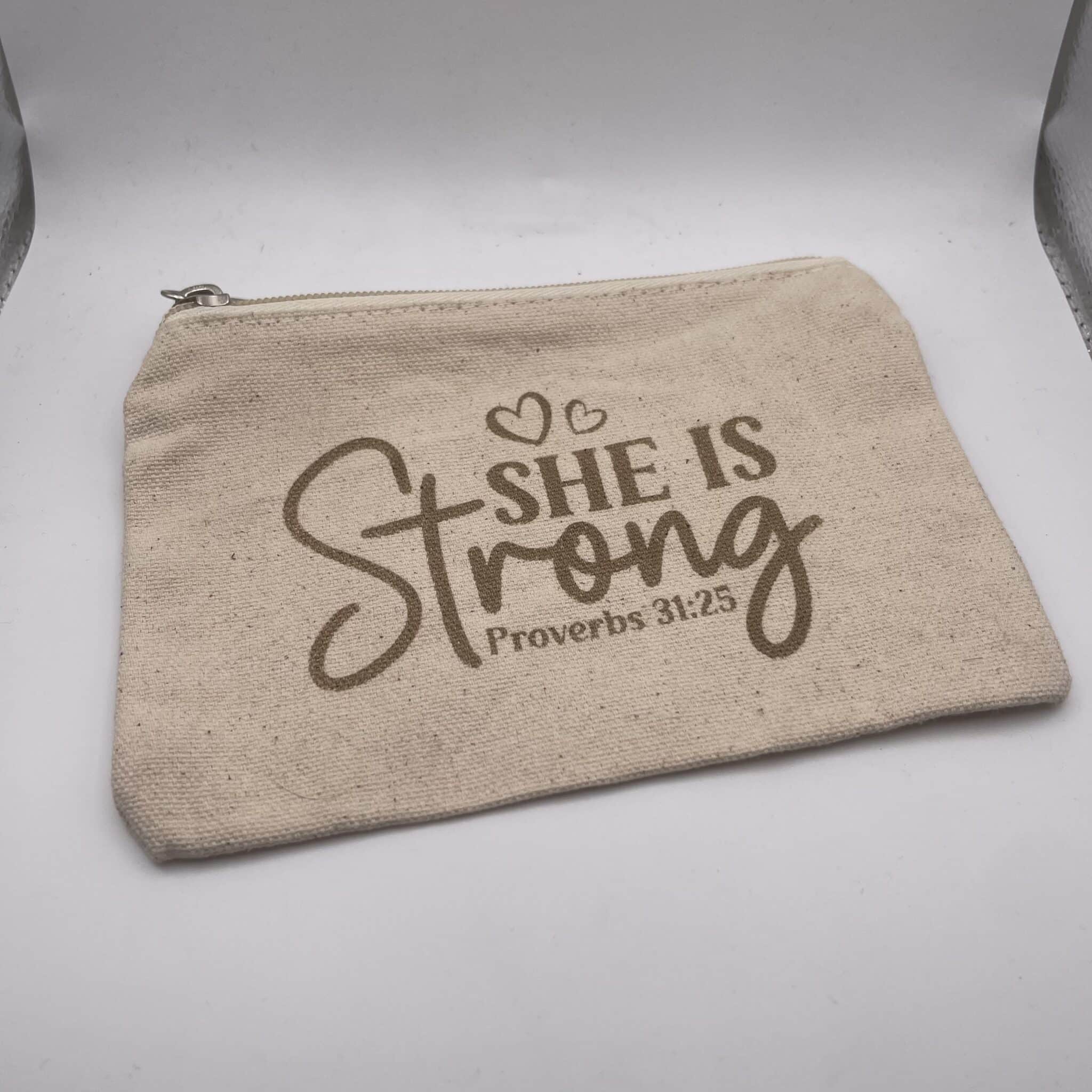 A Custom Engraved Natural Canvas Zipper Pouch in beige, featuring “She is Strong Proverbs 31:25” and two heart outlines printed in brown text on the front.