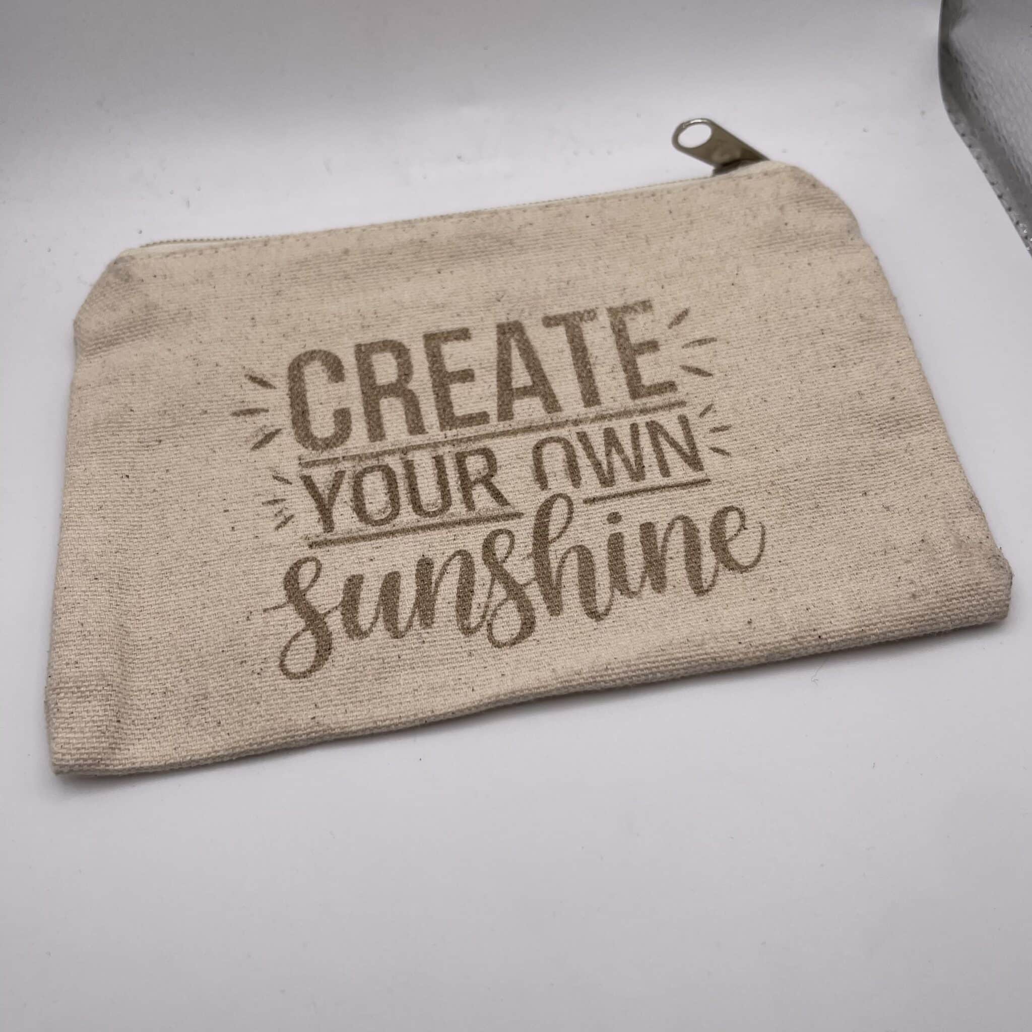 A Custom Engraved Natural Canvas Zipper Pouch in beige, featuring the phrase “CREATE YOUR OWN sunshine” in brown decorative lettering, displayed on a white surface.