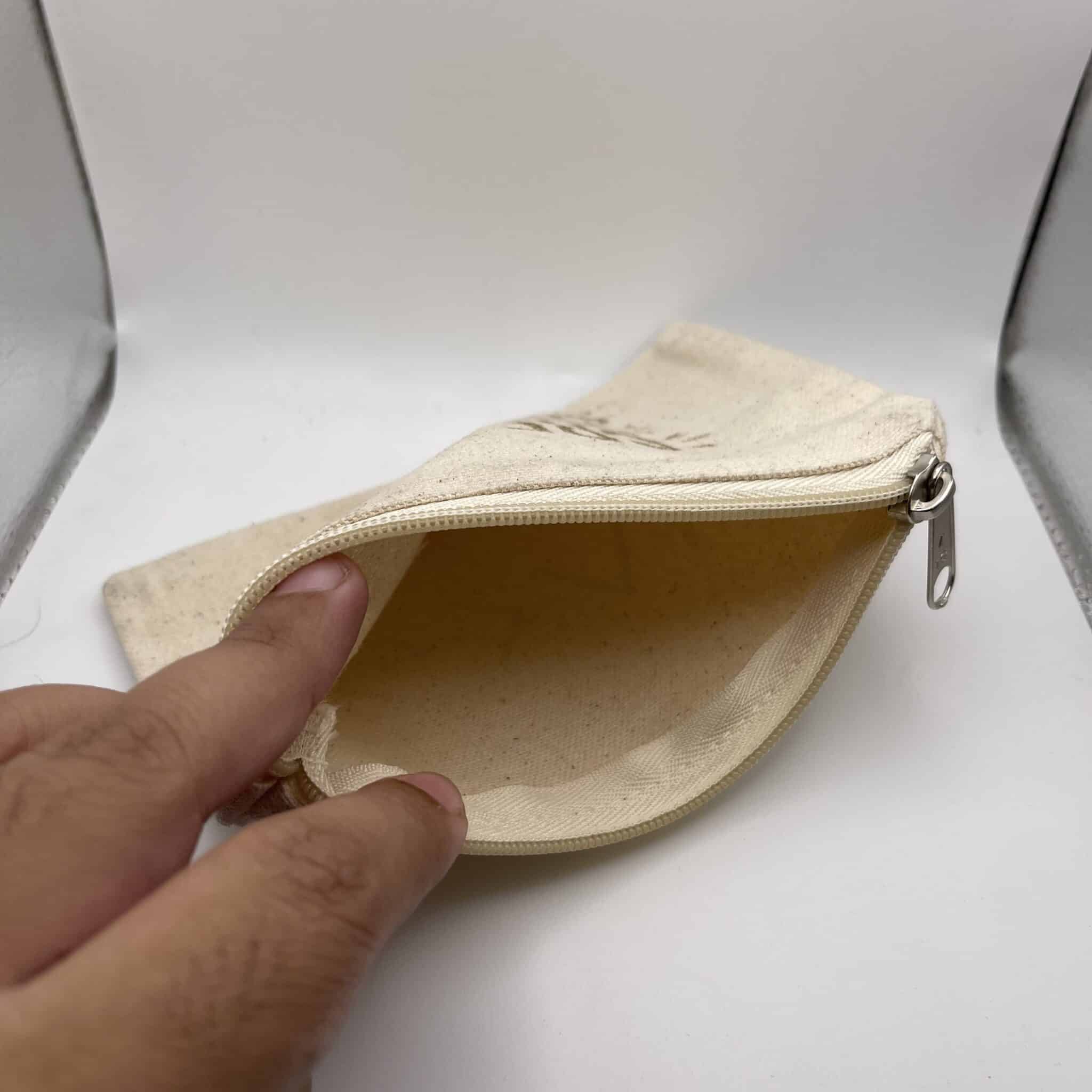 A hand holds open a Custom Engraved Natural Canvas Zipper Pouch, displaying its empty interior against a plain white background.