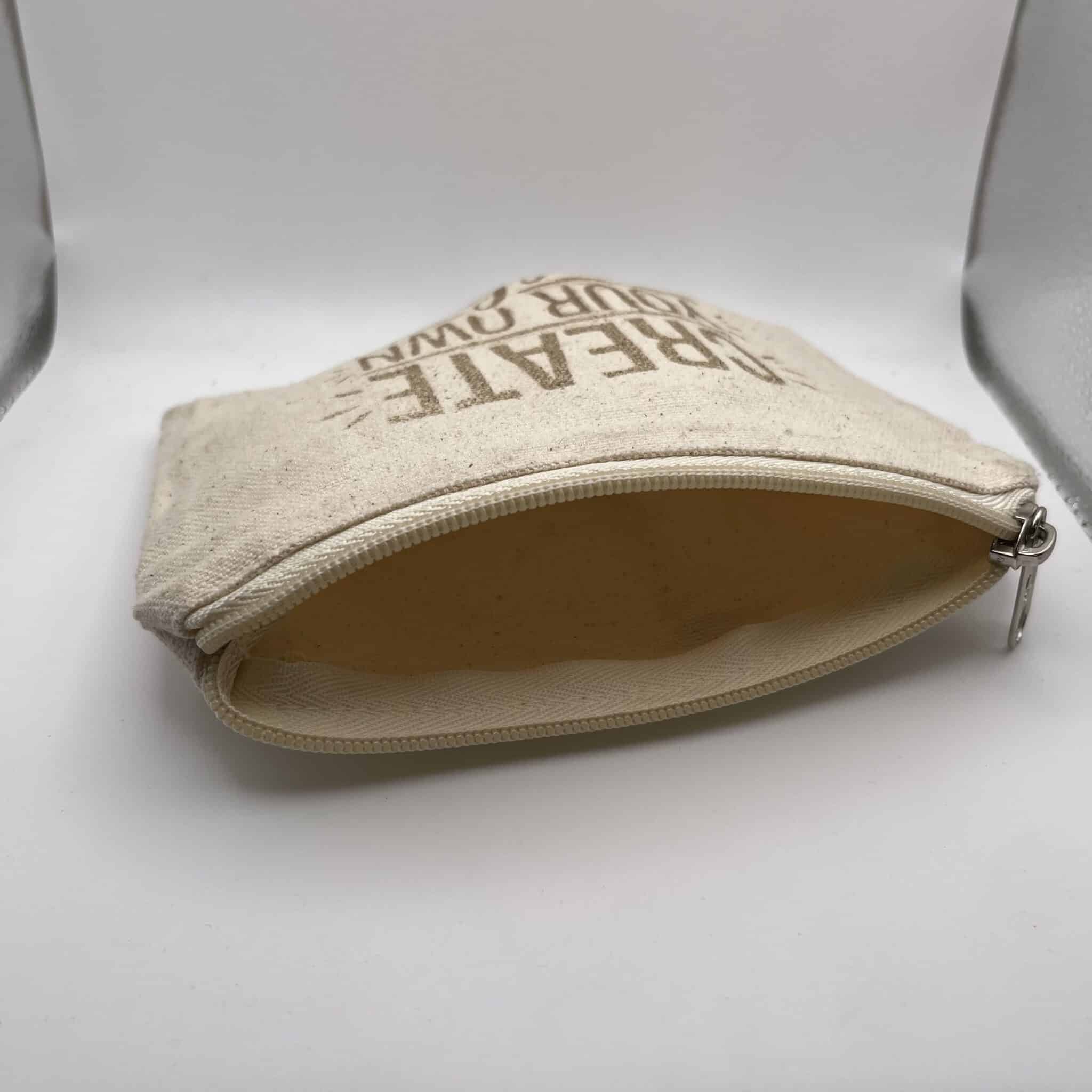 A Custom Engraved Natural Canvas Zipper Pouch in a small size and beige color is pictured unzipped and empty, with partially visible text on the front, lying on a white surface and light gray background.