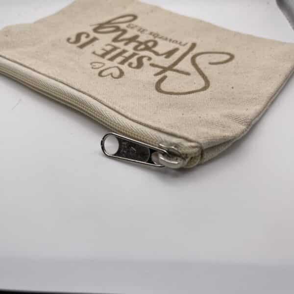 A Custom Engraved Natural Canvas Zipper Pouch in a small beige design rests on a white surface, featuring the phrase “SHE IS strong Proverbs 31:25” printed in brown on the fabric.