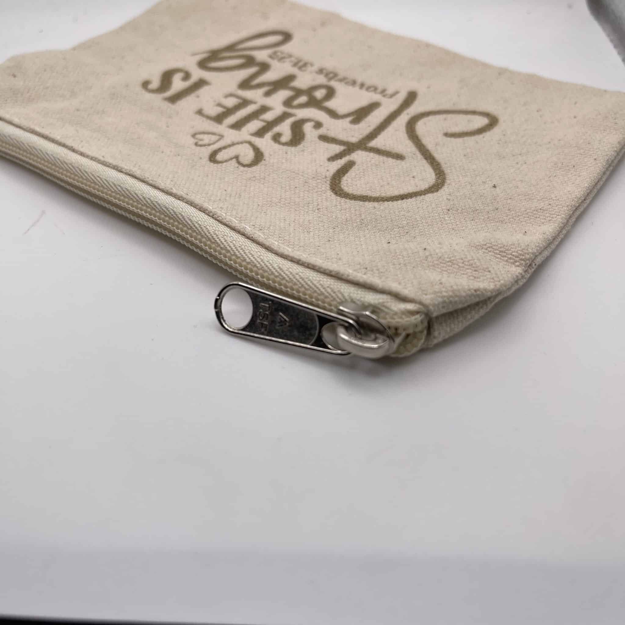 A Custom Engraved Natural Canvas Zipper Pouch in a small beige design rests on a white surface, featuring the phrase “SHE IS strong Proverbs 31:25” printed in brown on the fabric.