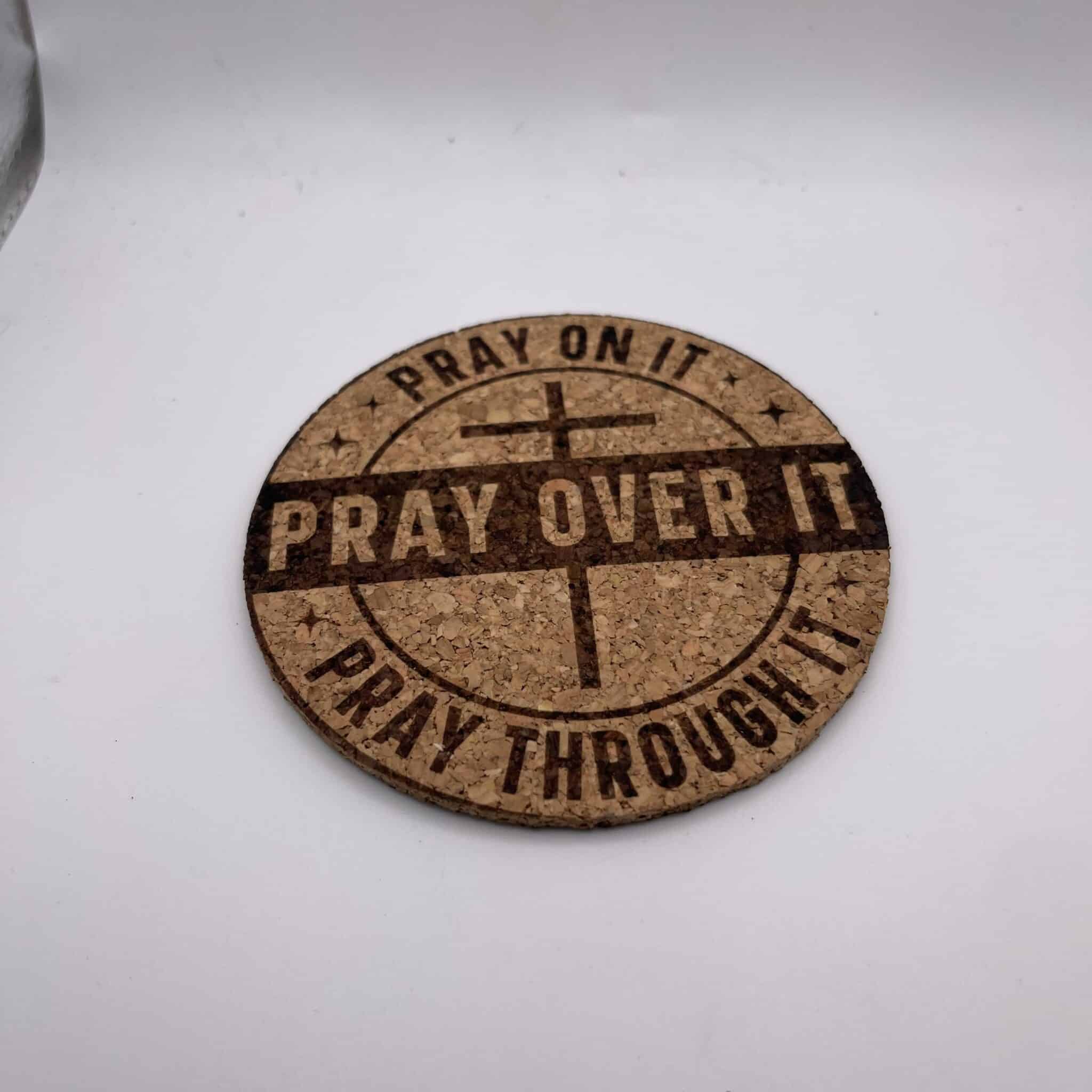A Circular 0.15″ Thick Cork Coaster engraved with a cross symbol and the phrases PRAY ON IT, PRAY OVER IT, and PRAY THROUGH IT rests on a white surface.