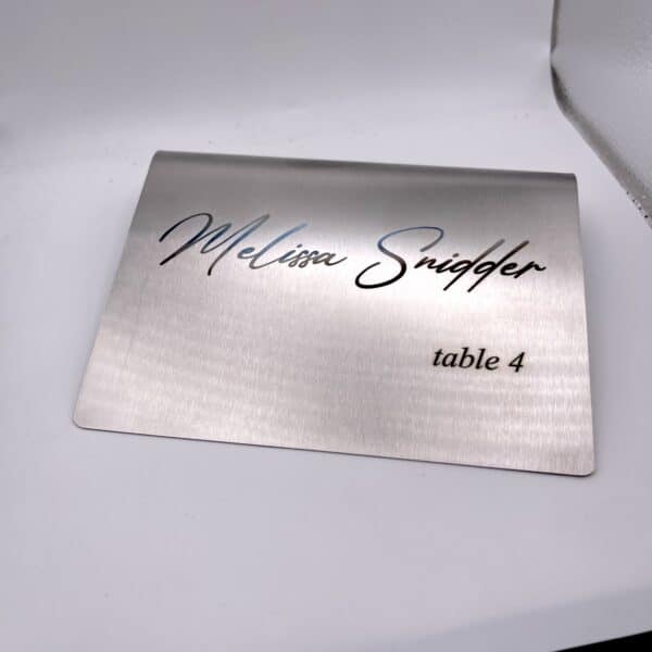 A Stainless Steal Place Holder Card displays “Melissa Snidger” in cursive and “table 4” in smaller print, set on a white surface.