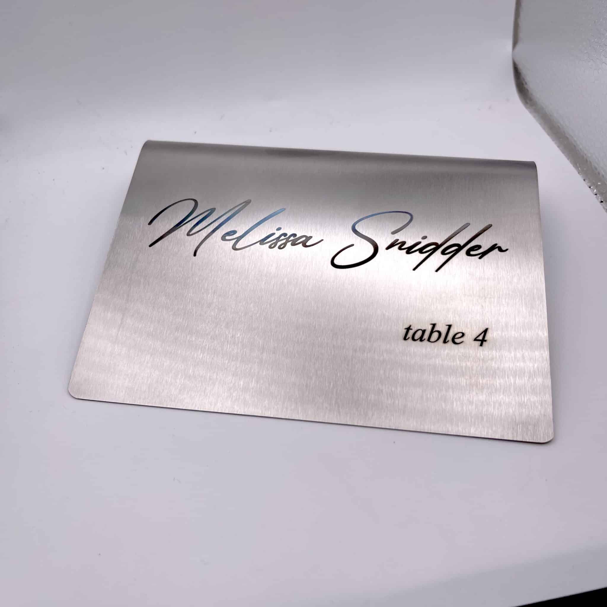 A Stainless Steal Place Holder Card displays “Melissa Snidger” in cursive and “table 4” in smaller print, set on a white surface.