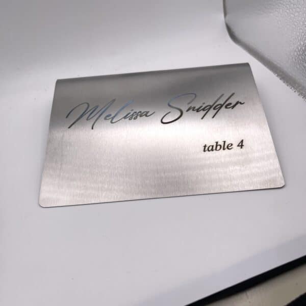 A Stainless Steel Place Holder Card displays “Melissa Snidger” in script and “table 4” in print, resting on a white surface beside a frosted glass item.