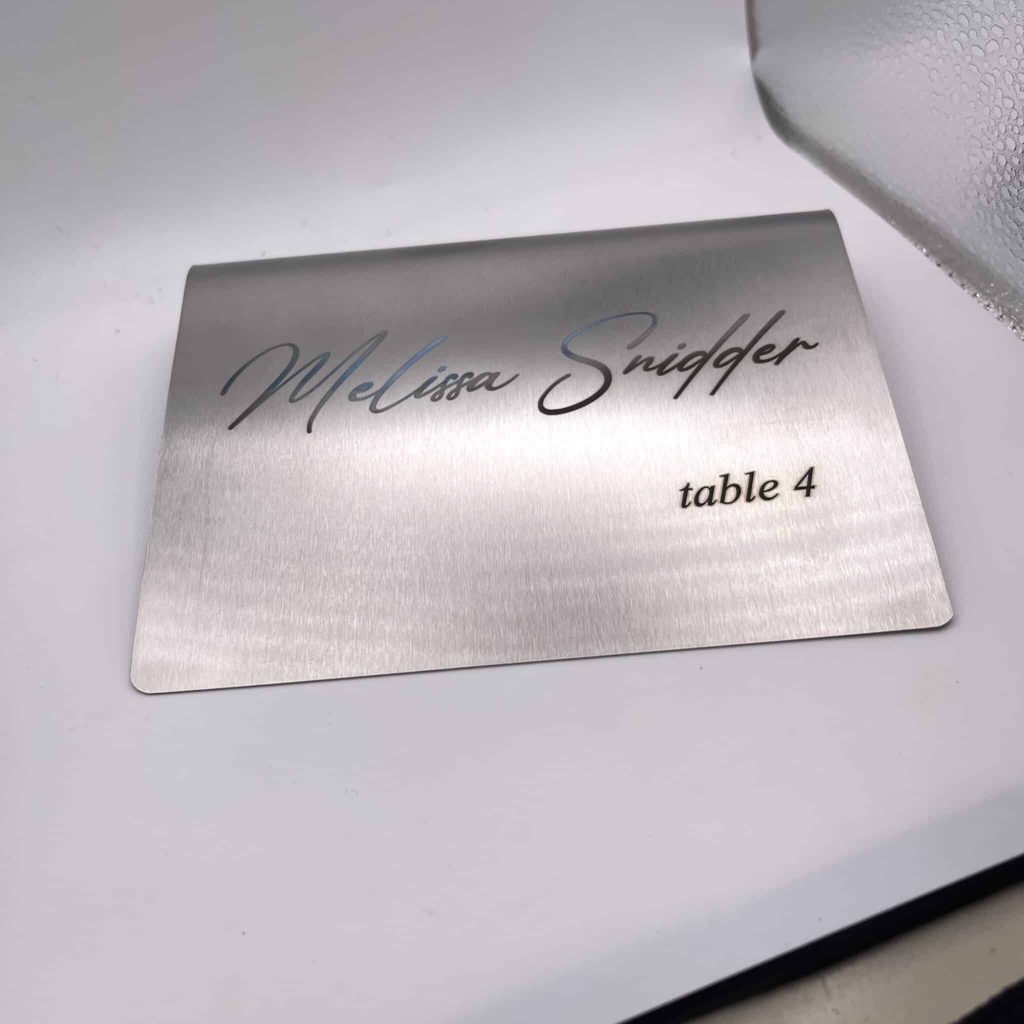 A Stainless Steel Place Holder Card displays “Melissa Snidger” in script and “table 4” in print, resting on a white surface beside a frosted glass item.