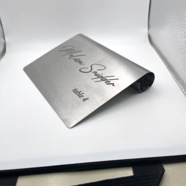 A Stainless Steal Place Holder Card, designed as a desk name plaque, displays “Melissa Snidger” in script and “table 4” in print. The mini sign is bent to stand upright and photographed against a plain white background.