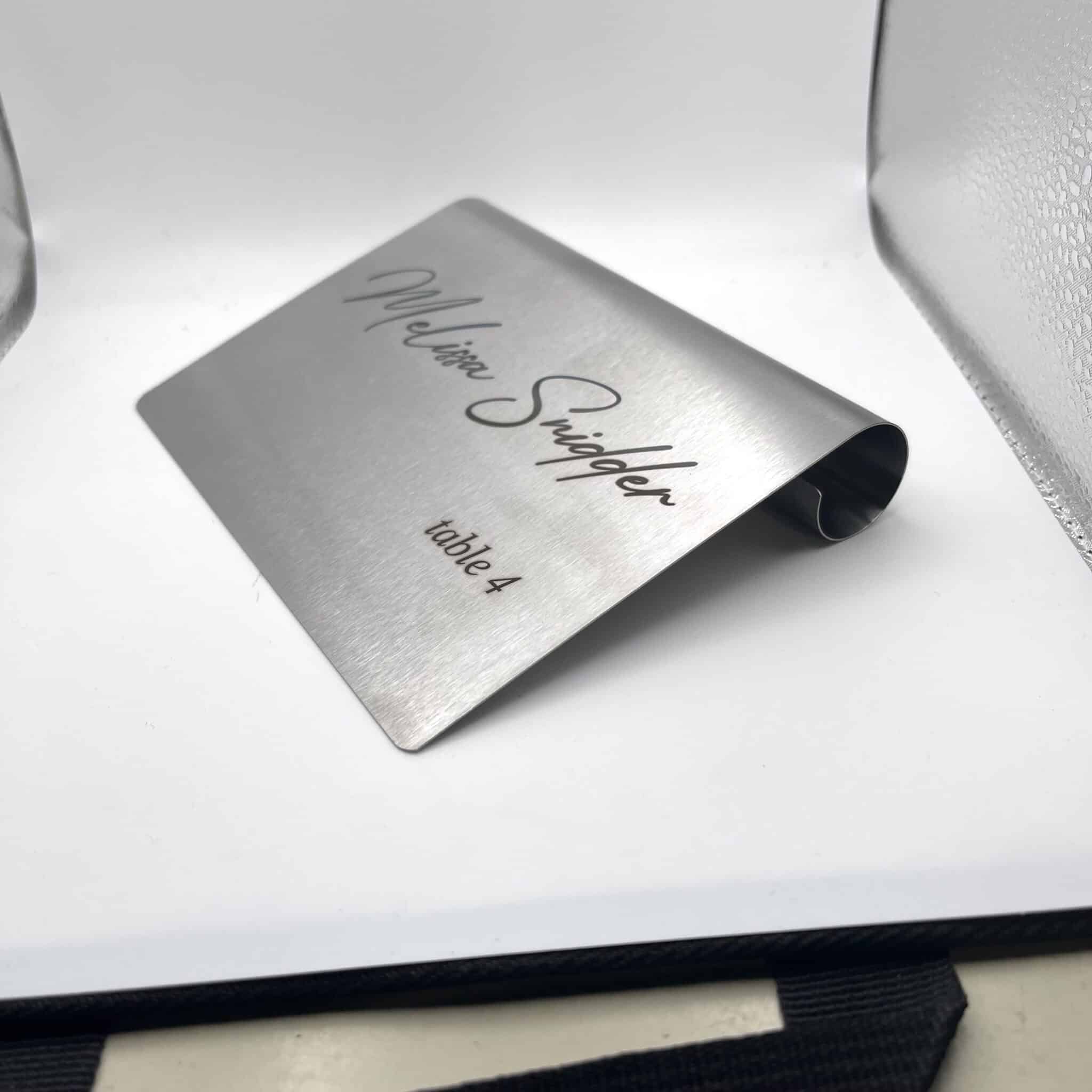 A Stainless Steal Place Holder Card, designed as a desk name plaque, displays “Melissa Snidger” in script and “table 4” in print. The mini sign is bent to stand upright and photographed against a plain white background.