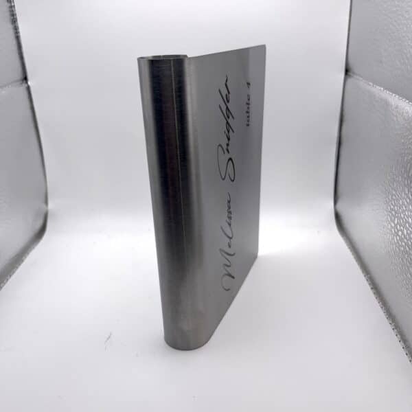 A Stainless Steel Place Holder Card with “McClure Snuggler table 4” engraved, standing upright against a white and silver reflective background.