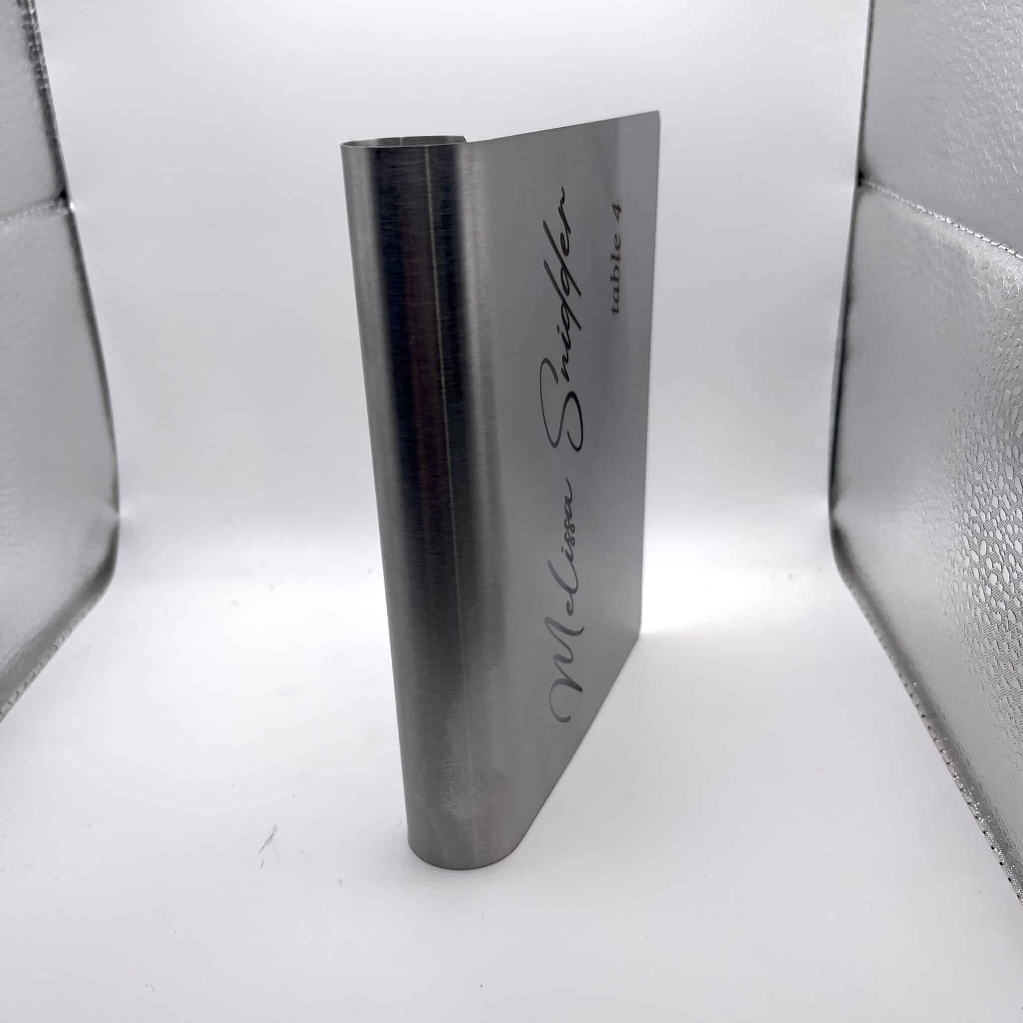 A Stainless Steel Place Holder Card with “McClure Snuggler table 4” engraved, standing upright against a white and silver reflective background.