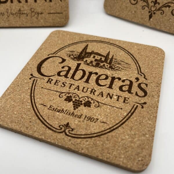A Square 0.20″ Thick Cork Coaster engraved with “Cabreras Restaurante,” featuring an illustration of a villa with trees and the text “Established 1907” on a banner; additional similar coasters appear partially visible in the background.
