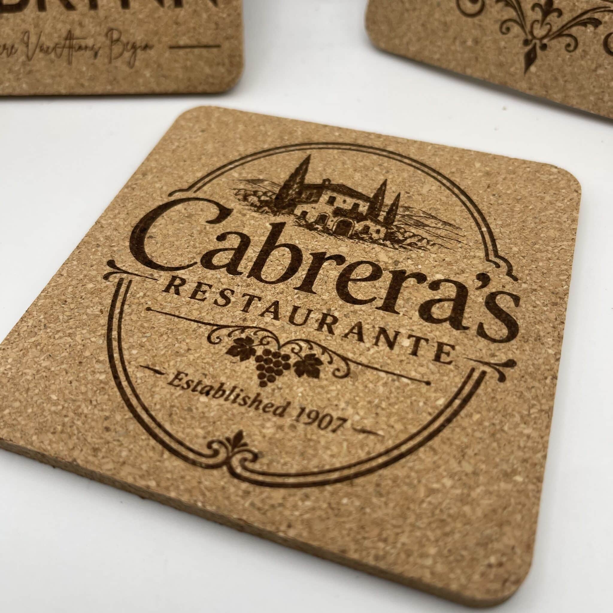A Square 0.20″ Thick Cork Coaster engraved with “Cabreras Restaurante,” featuring an illustration of a villa with trees and the text “Established 1907” on a banner; additional similar coasters appear partially visible in the background.