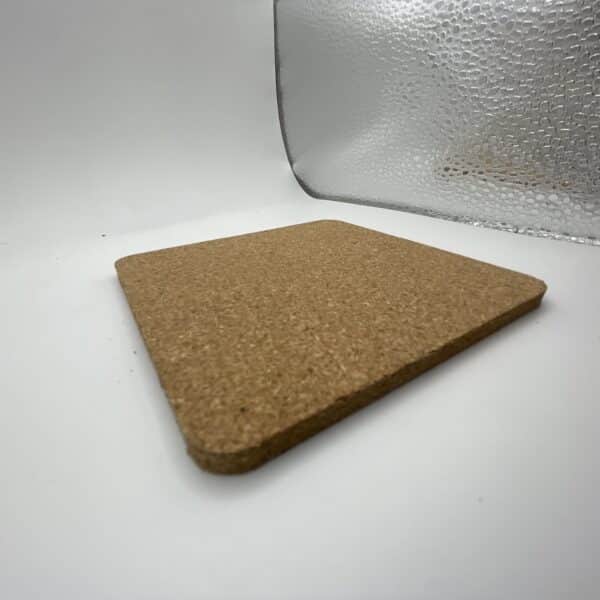A Square 0.20″ Thick Cork Coaster rests on a white surface, with a textured, transparent glass object placed behind it.