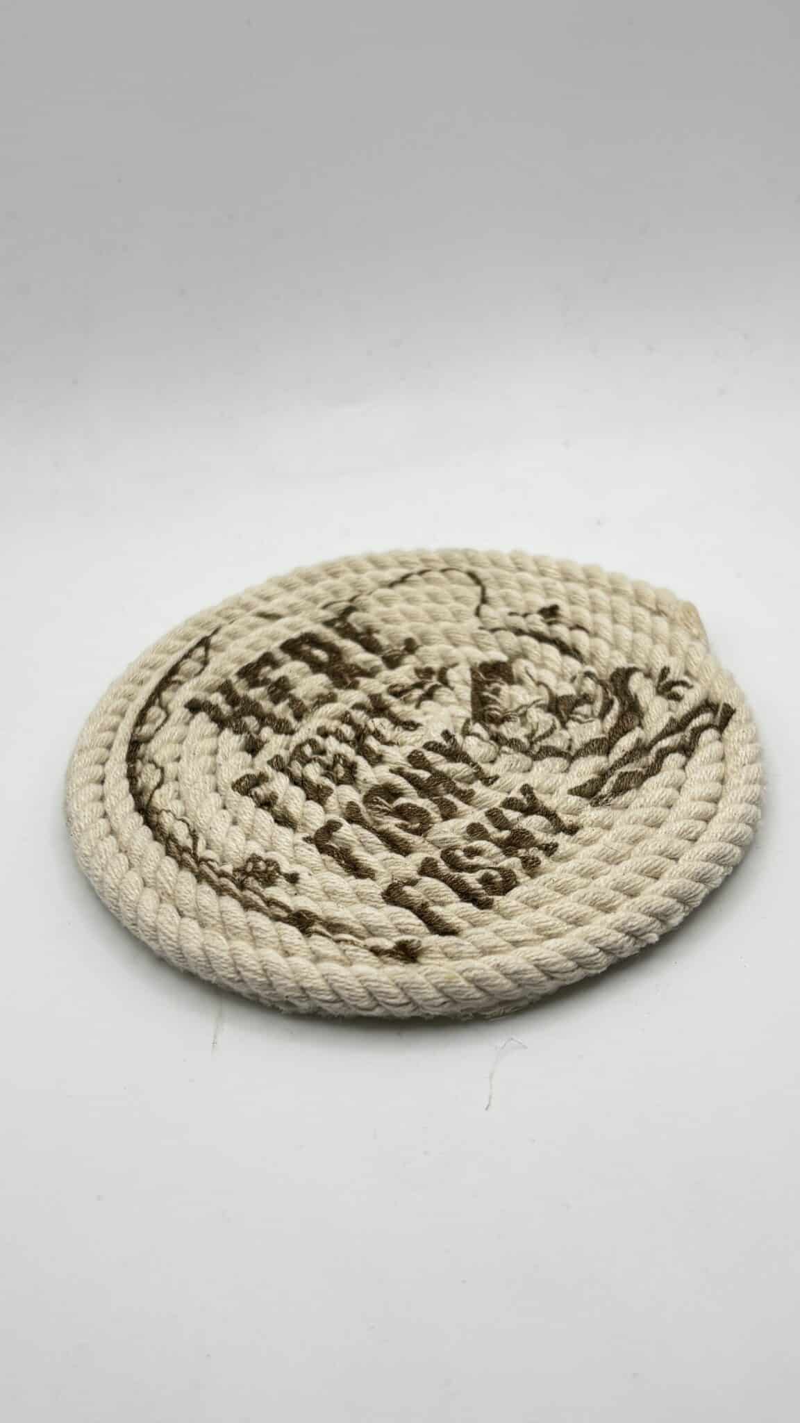 A round, coiled rope mat with brown text and a design on its surface, placed on a plain white background.