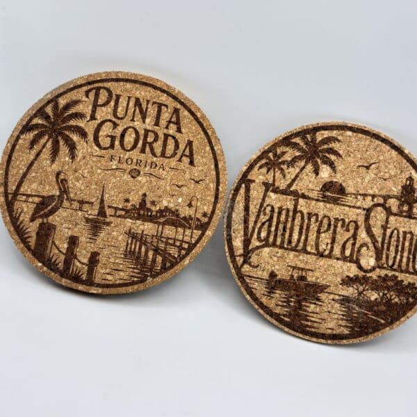 A pair of Circular 0.15″ Thick Cork Coasters with engraved designs is shown upright on a white background. One coaster features the text “Punta Gorda Florida” alongside palm trees and a pelican, while the other displays “Manbrera Sones” with a bridge, trees, and water scene.