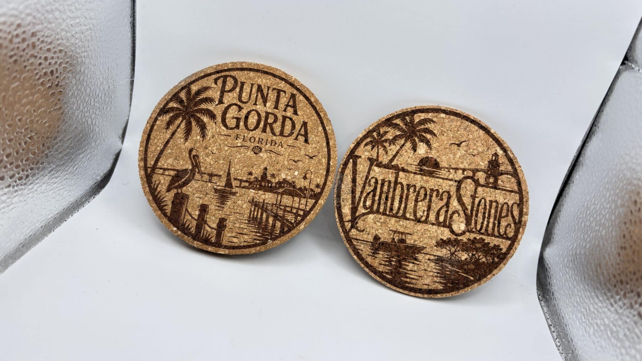 A pair of Circular 0.15″ Thick Cork Coasters with engraved designs is shown upright on a white background. One coaster features the text “Punta Gorda Florida” alongside palm trees and a pelican, while the other displays “Manbrera Sones” with a bridge, trees, and water scene.