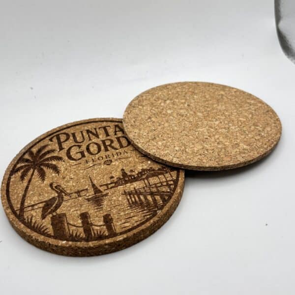 Two Circular 0.15″ Thick Cork Coasters rest on a white surface—one engraved with a Punta Gorda, Florida beach scene featuring palm trees and a pier, while the other is blank.