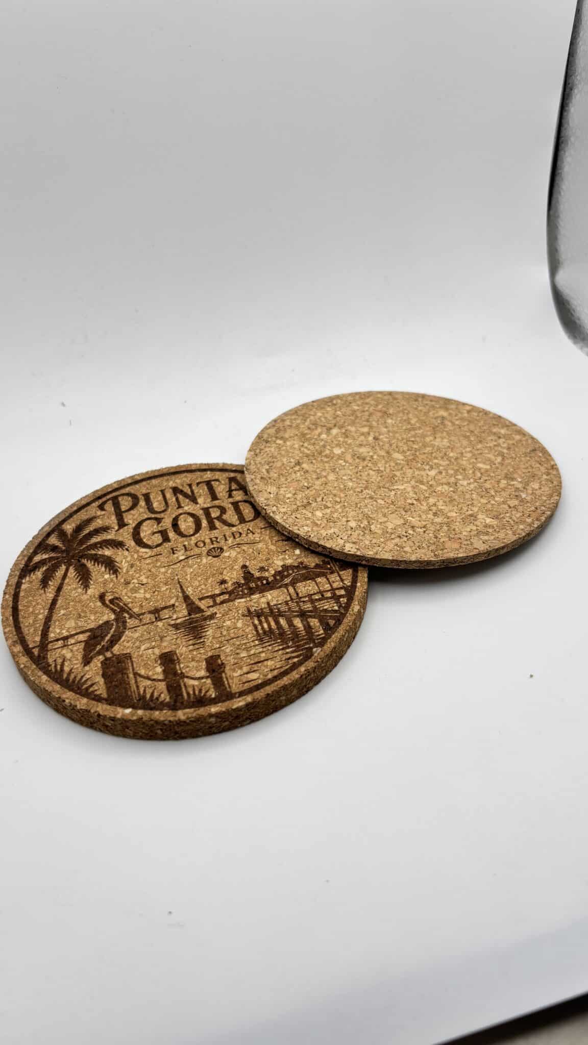 Two Circular 0.15″ Thick Cork Coasters rest on a white surface—one engraved with a Punta Gorda, Florida beach scene featuring palm trees and a pier, while the other is blank.