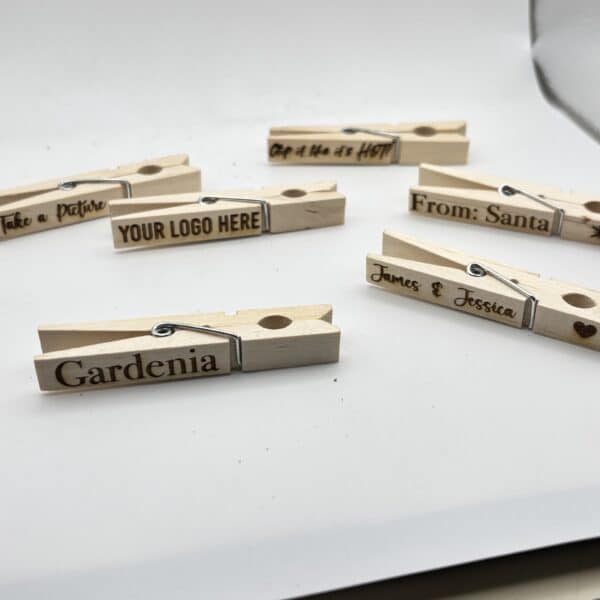 Six wooden clothespins with various engraved messages, including Gardenia, From: Santa, and YOUR LOGO HERE, are displayed on a white surface with a reflective background.