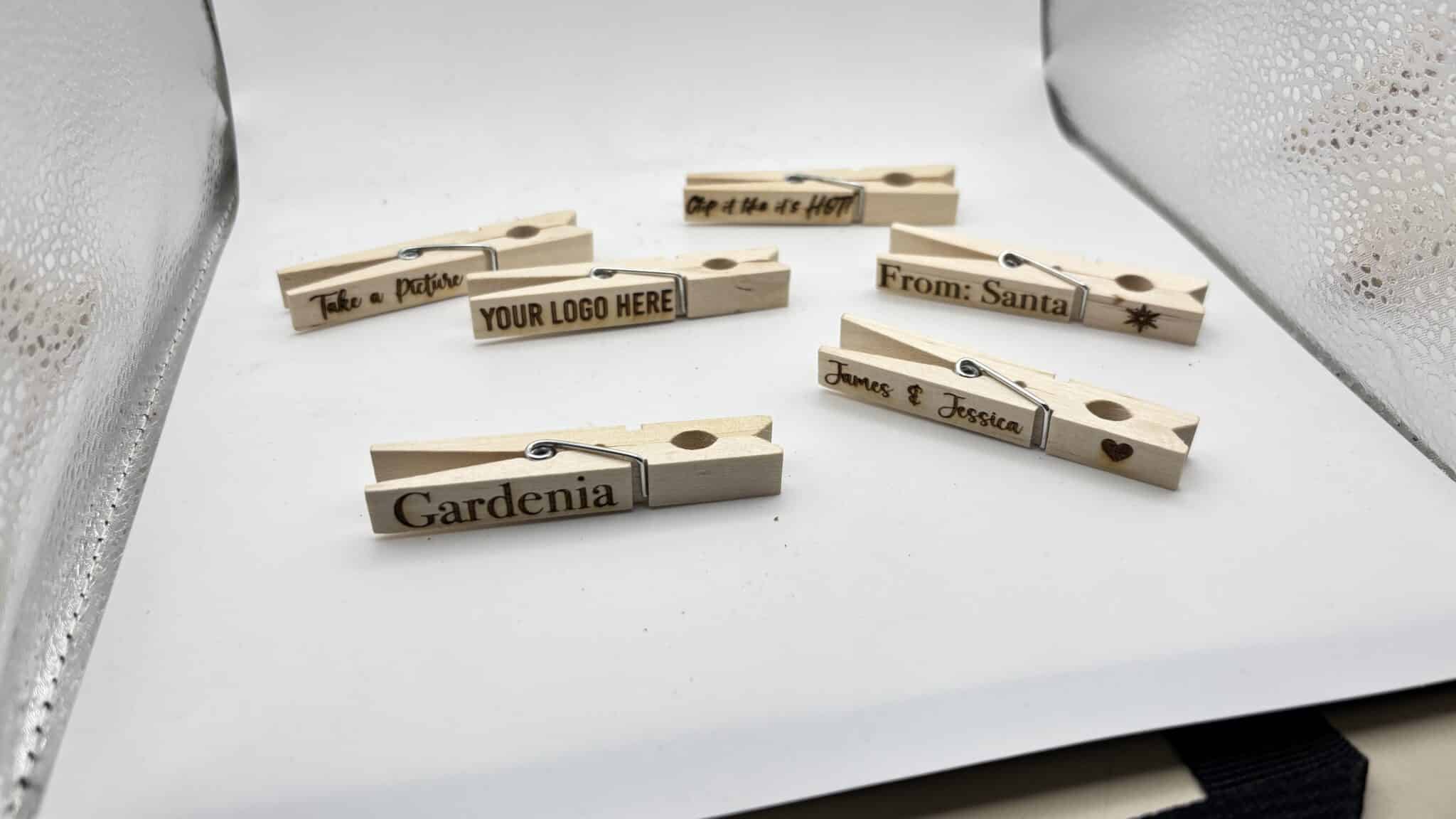 Six wooden clothespins with various engraved messages, including Gardenia, From: Santa, and YOUR LOGO HERE, are displayed on a white surface with a reflective background.