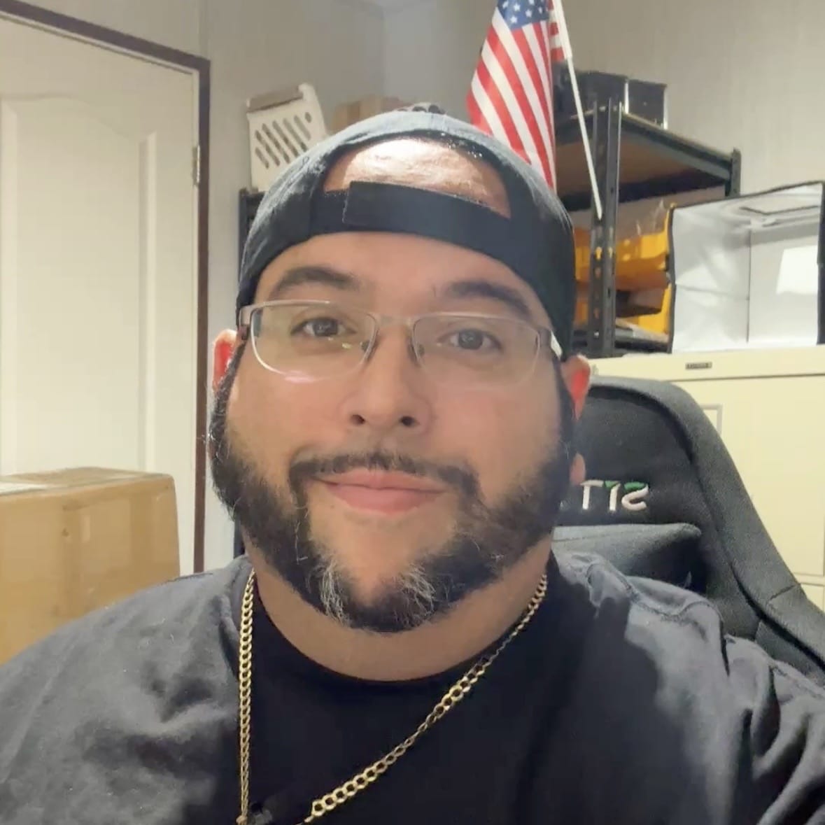 Screenshot A man with glasses and a black cap worn backward sits in an office with shelves, a box, and a U.S. flag in the background. He wears a black shirt and gold chain, and smiles at the camera.